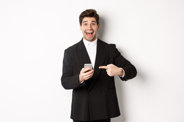 Immagine di un uomo d'affari allegro che sembra stupito, indicando il telefono cellulare, in piedi in tuta su sfondo bianco