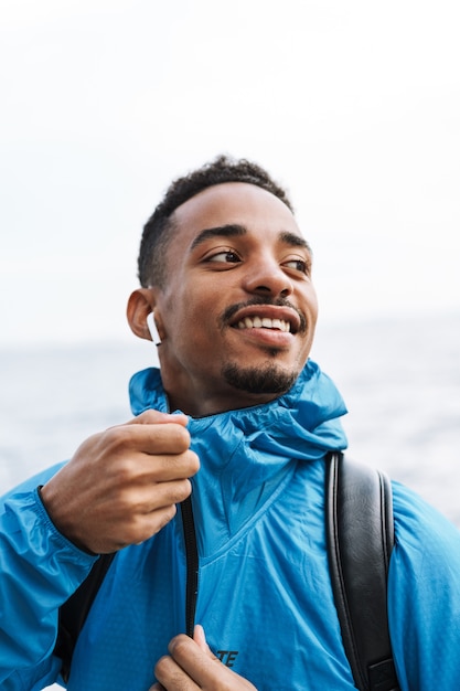 Immagine di un uomo di sport africano giovane felice bello all'aperto in spiaggia mare a piedi con la borsa ascoltando musica con gli auricolari.