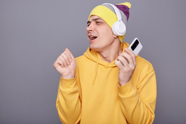 Immagine di un uomo divertente e positivo che indossa un berretto e una felpa con cappuccio casual gialla in piedi isolato su sfondo grigio che ascolta musica o radio in cuffia che canta tenendo il cellulare