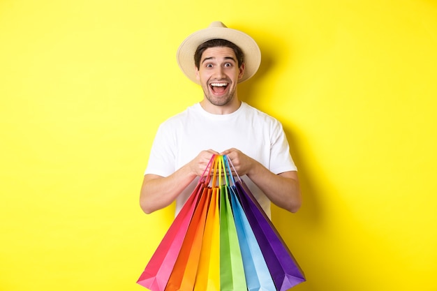 Immagine di un uomo felice che fa shopping in vacanza, tiene in mano sacchetti di carta e sorride, in piedi su sfondo giallo