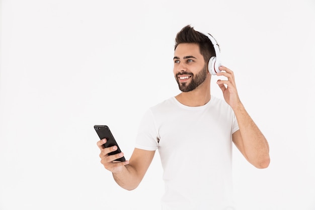 Immagine di un uomo soddisfatto con la barba lunga che ascolta musica sul cellulare e sulle cuffie isolate su un muro bianco