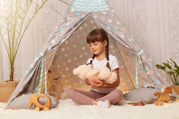 Immagine di una bambina affascinante che indossa una maglietta bianca seduta sul pavimento a wigwam e gioca con l'orsacchiotto che tiene il giocattolo in mano mettendo il suo giocattolo preferito a dormire