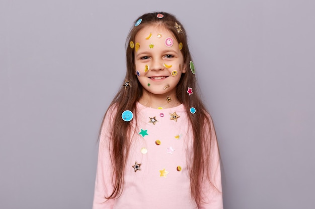 Immagine di una bambina dai capelli castani piuttosto carina e sorridente ricoperta di adesivi che guarda l'obbiettivo con espressione soddisfatta essendo di buon umore in posa isolata su sfondo grigio