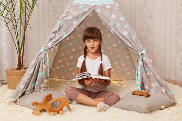 Immagine di una bambina scioccata e stupita con trecce che indossa una maglietta bianca e leggins che legge un libro guardando la fotocamera con la bocca aperta che è impressionata da una fiaba interessante