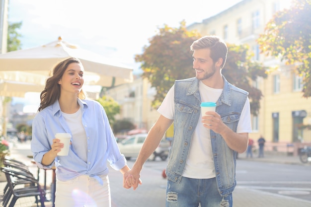 Immagine di una bella coppia felice in abiti estivi che sorridono e si tengono per mano mentre camminano per le strade della città.