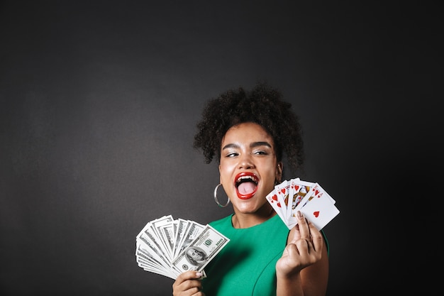 Immagine di una bella donna africana in posa isolata sopra il muro nero che tiene soldi e carte da gioco.