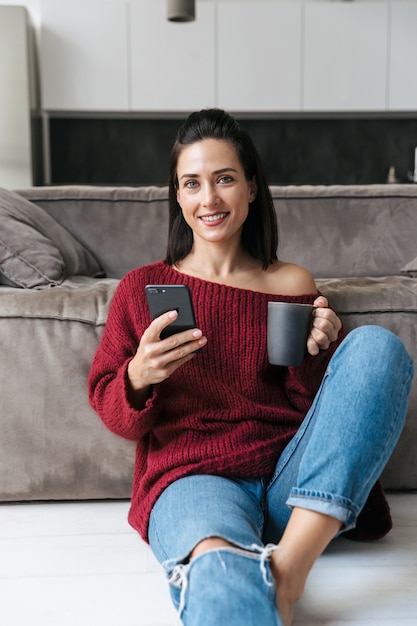 Immagine di una bella donna al chiuso in casa vicino al divano utilizzando il telefono cellulare bere caffè.