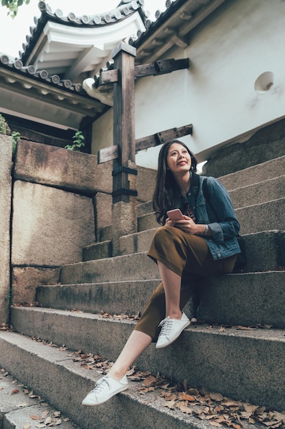 Immagine di una bella donna asiatica da viaggio alla moda seduta sulle scale che tiene il telefono cellulare che guarda da parte mentre si visita il castello di osaka. la ragazza si rilassa sul gradino usando il monumento all'aperto dello smartphone
