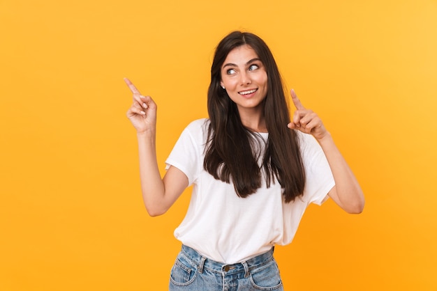 Immagine di una bella donna bruna con i capelli lunghi che sorride e punta il dito contro copyspace isolato su un muro giallo