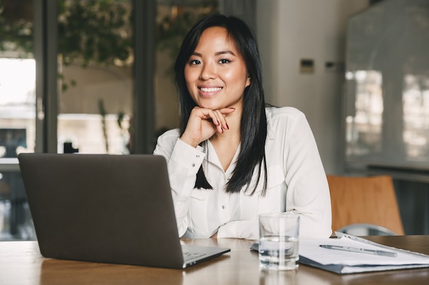 Immagine di una bella donna d'affari asiatici che indossa una camicia bianca, sorridente mentre era seduto al tavolo in ufficio, durante il lavoro sul computer portatile