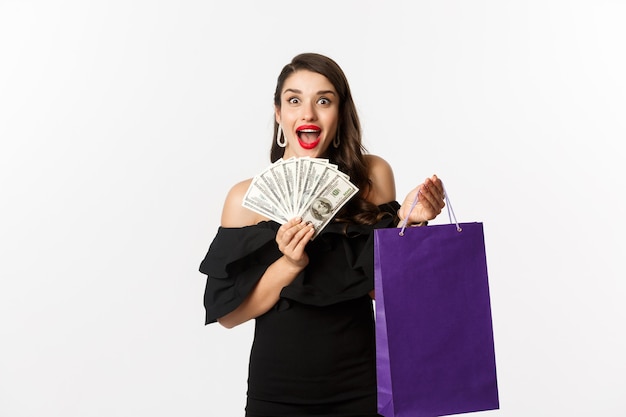 Immagine di una bella donna eccitata in abito nero che fa shopping, tiene in mano borsa e dollari, in piedi su sfondo bianco.