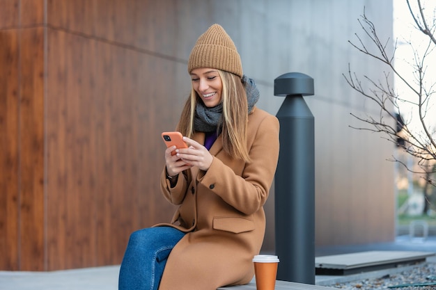 Immagine di una bella donna elegante seduta su una panchina di strada con le gambe incrociate in una giornata di sole e con in mano un telefono cellulare