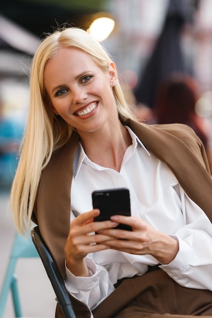 Immagine di una bella donna sorridente che indossa una giacca che digita sul cellulare mentre è seduta al bar all'aperto