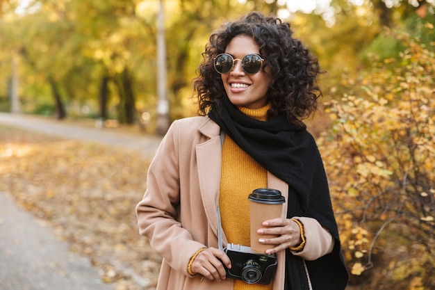 Immagine di una bella giovane donna africana che cammina all'aperto in un parco primaverile bevendo caffè.