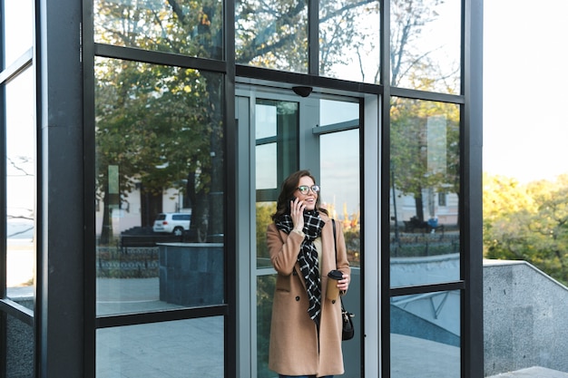 Immagine di una bella giovane donna felice all'aperto camminando per strada a bere caffè parlando con il telefono cellulare.