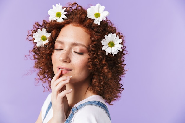 Immagine di una bella ragazza rossa felice riccia in posa isolata sul muro viola con fiori nei capelli.