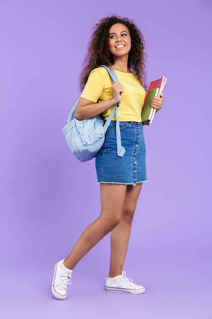 Immagine di una bella studentessa afroamericana con i capelli ricci che tiene quaderni isolati su un muro viola