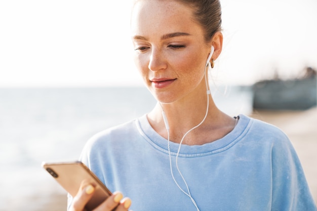 Immagine di una bellissima giovane donna fitness concentrata all'aperto sulla spiaggia utilizzando la musica d'ascolto del telefono cellulare con gli auricolari.