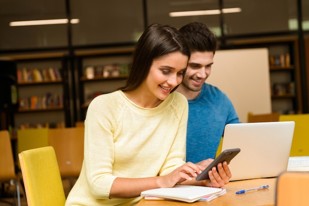 Immagine di una coppia di amici giovani studenti in biblioteca a fare i compiti studiando leggere e utilizzando computer portatile e telefono cellulare.