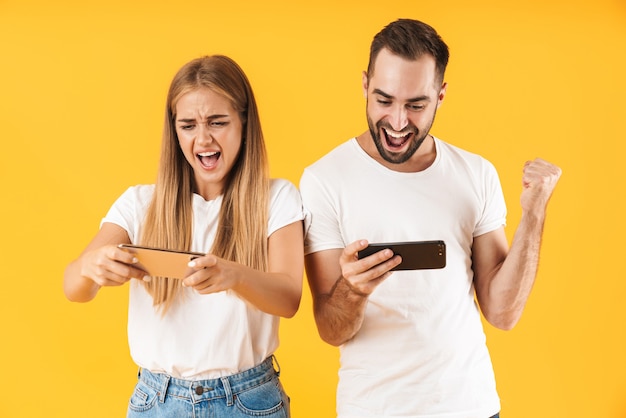 Immagine di una coppia eccitata uomo e donna in magliette di base che giocano ai videogiochi su smartphone insieme isolati su un muro giallo
