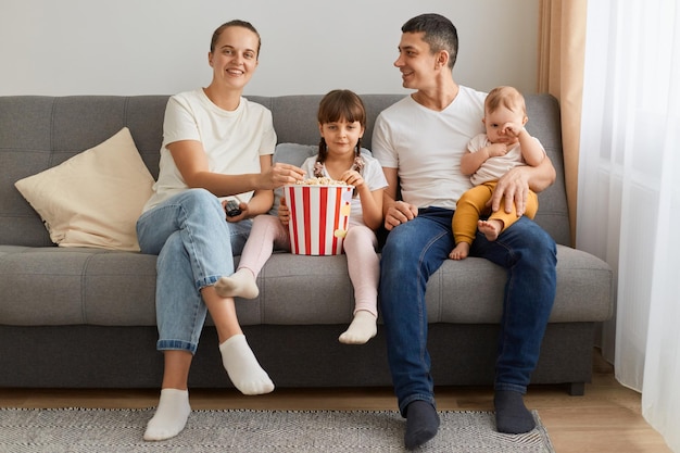 Immagine di una coppia ottimista con i loro figli seduti sul divano a casa in soggiorno e guardando un interessante programma televisivo o film che esprimono emozioni positive e sorridono