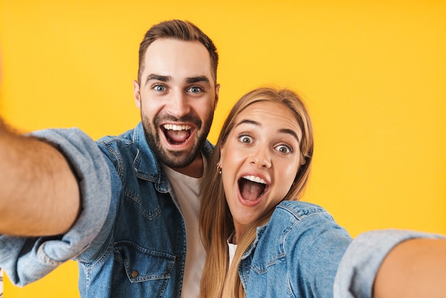 Immagine di una coppia stupita uomo e donna in abiti di jeans che urlano mentre scattano foto selfie isolate su un muro giallo