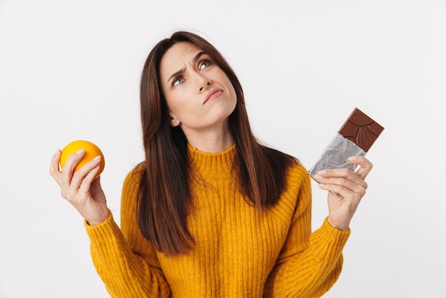 Immagine di una donna adulta bruna dubbiosa che esita mentre tiene in mano una barra di cioccolato e arancia isolata su bianco