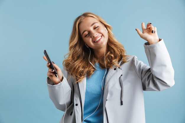 Immagine di una donna affascinante 20s che indossa un impermeabile che tiene il telefono cellulare e ascolta la musica tramite gli auricolari