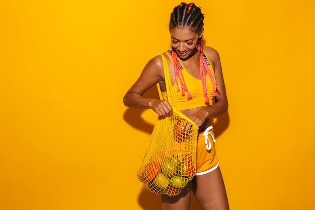 Immagine di una donna afroamericana positiva che indossa trecce afro sorridenti e porta una borsa a rete con frutti isolati su un muro giallo