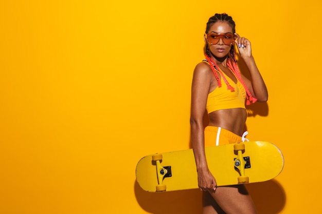 Immagine di una donna afroamericana seria che indossa occhiali da sole che tiene in mano lo skateboard e guarda da parte isolata su un muro giallo