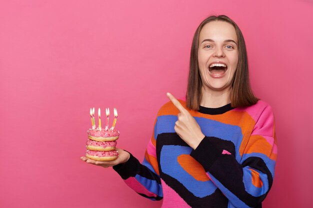 Immagine di una donna attraente e felice con i capelli castani che indossa un maglione elegante che punta verso l'area pubblicitaria tenendo una ciambella con candele isolate su sfondo rosa