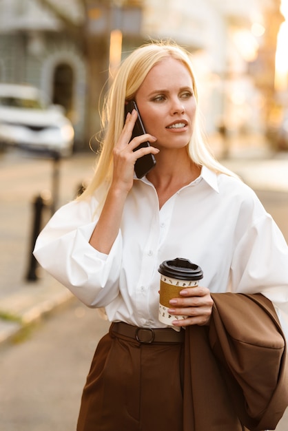 Immagine di una donna bionda seria che indossa una camicia bianca che parla al cellulare e beve caffè da asporto mentre si cammina per la strada della città