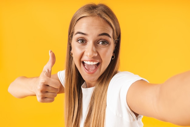 Immagine di una donna carina in abiti semplici che sorride e mostra il pollice in su mentre scatta una foto selfie isolata su un muro giallo