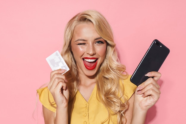 Immagine di una donna caucasica con lunghi capelli biondi che sorride mentre tiene il cellulare e la carta di credito isolati su un muro rosa