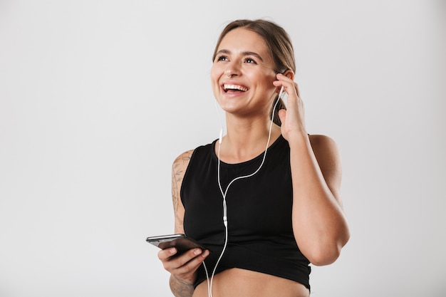 Immagine di una donna caucasica in abiti sportivi che tiene in mano uno smartphone e ascolta musica con gli auricolari isolati su un muro bianco