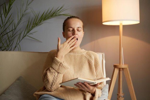 Immagine di una donna caucasica stanca, annoiata e assonnata con un'acconciatura da panino che indossa un maglione beige che legge un libro che sbadiglia coprendo la bocca con il palmo seduto sul divano nel soggiorno