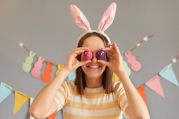 Immagine di una donna dai capelli castani sorridente e felice che indossa orecchie di coniglio coprendosi gli occhi con uova di Pasqua colorate isolate su sfondo grigio decorato sorridendo alla telecamera che celebra le vacanze