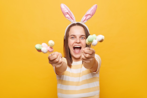 Immagine di una donna eccitata festiva positiva vestita di orecchie di coniglio che tiene fuori mostrando la torta di Pasqua schiocca isolata su sfondo giallo urlando con faccia positiva godendosi le vacanze