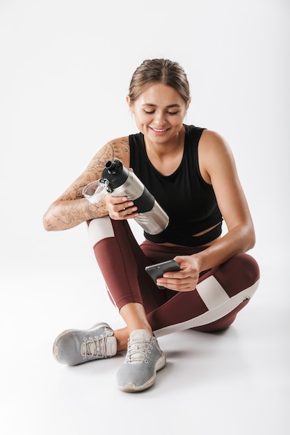 Immagine di una donna felice in abiti sportivi che tiene cellulare e bottiglia d'acqua mentre è seduta sul pavimento isolata su un muro bianco