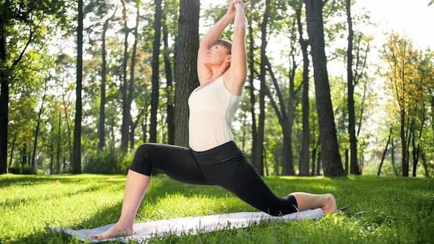 Immagine di una donna felice sorridente di mezza età che medita e fa esercizi di yoga sull'erba della foresta. Donna che si prende cura della sua salute fisica e mentale mentre pratica fitness e si estende al parco