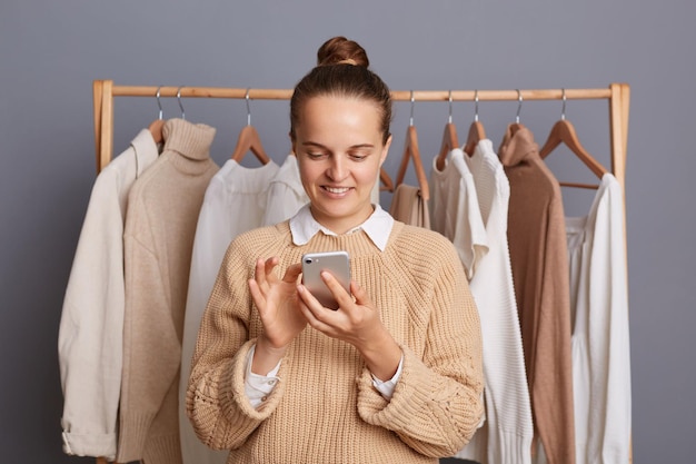 Immagine di una donna positiva concentrata che indossa un maglione beige in piedi contro il muro grigio con vestiti appesi nell'armadio su rack in posa con il telefono cellulare utilizzando lo smartphone