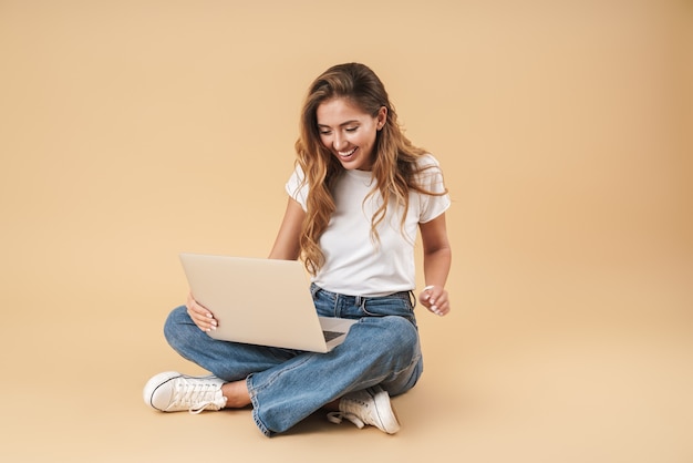 Immagine di una donna sorridente che indossa abiti casual usando un laptop argento mentre è seduta sul pavimento con le gambe incrociate isolate su un muro beige