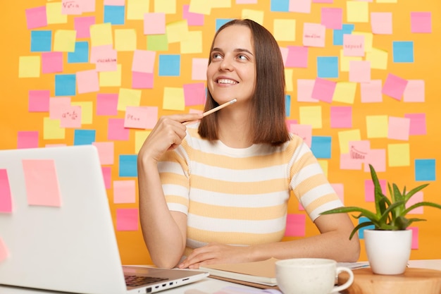 Immagine di una donna sorridente che sogna pensierosa con i capelli castani che indossa una maglietta seduta al tavolo in posa contro il muro giallo con piccole note adesive si siede davanti al laptop e pensa a qualcosa