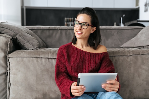 Immagine di una donna straordinaria al chiuso in casa vicino al divano utilizzando il computer tablet.