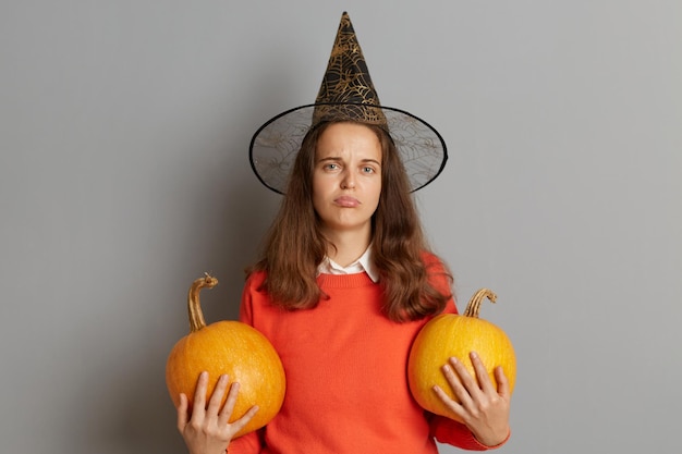 Immagine di una donna triste sconvolta che indossa un cappello da strega in piedi con due zucche arancioni isolate su sfondo grigio che celebra Halloween di cattivo umore esprimendo dolore e tristezza