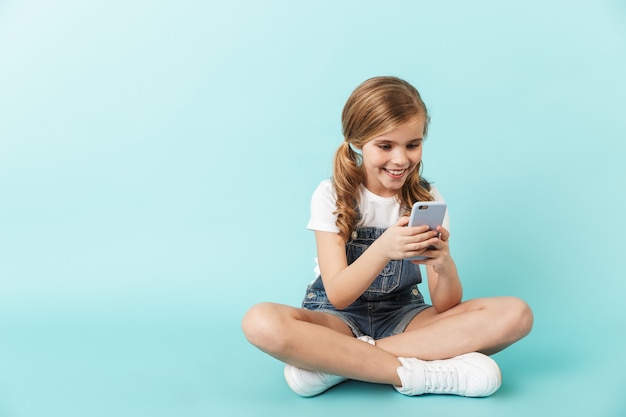 Immagine di una giovane bambina abbastanza felice in posa isolata su un telefono cellulare blu che usa il muro in chat.