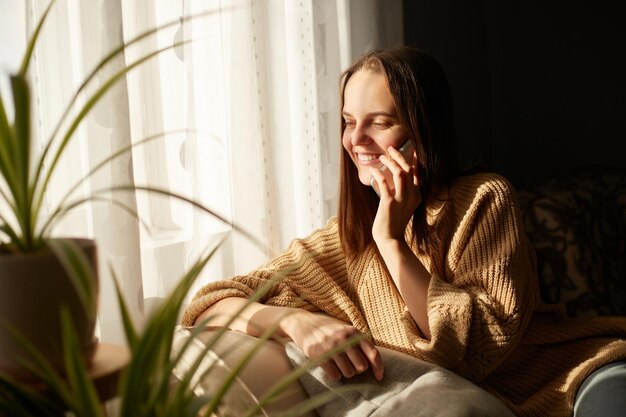 Immagine di una giovane bella donna che parla sullo smartphone con un'espressione felice a casa vicino alla finestra alla luce del sole che distoglie lo sguardo con un sorriso ascoltando notizie piacevoli o ha una conversazione romantica