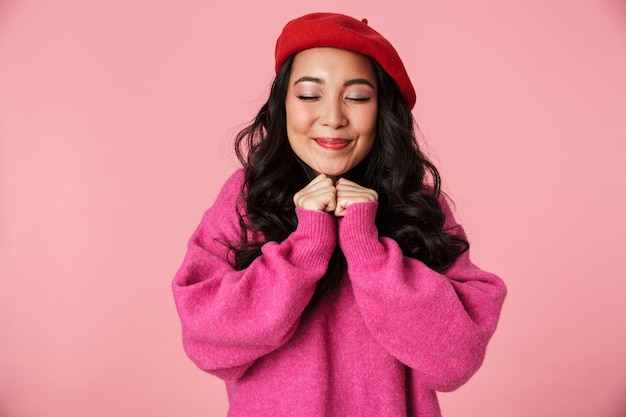 Immagine di una giovane bella ragazza asiatica che indossa un berretto che sorride e stringe i pugni al petto