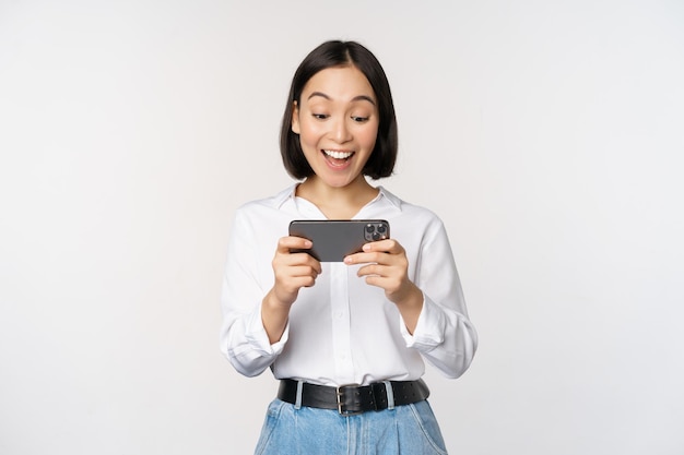 Immagine di una giovane donna asiatica che guarda sull'app per smartphone con in mano il telefono cellulare e guarda lo schermo ridendo e sorridendo in piedi su sfondo bianco