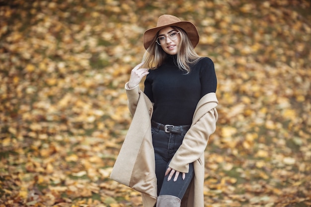 Immagine di una giovane donna attraente vestita con un cappotto e un cappello di feltro sullo sfondo delle foglie cadute nel parco in autunno